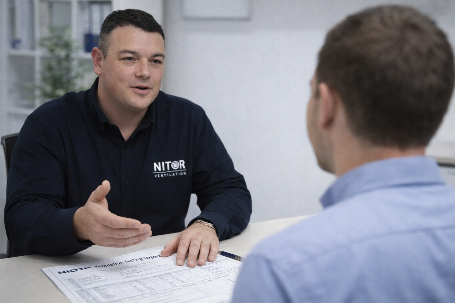 Man in dark shirt gestures, talking to another man at a desk, document visible.