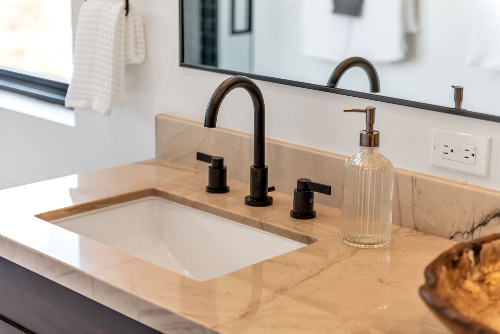 Bathroom sink with black faucet on marble countertop, glass soap dispenser, and mirror.