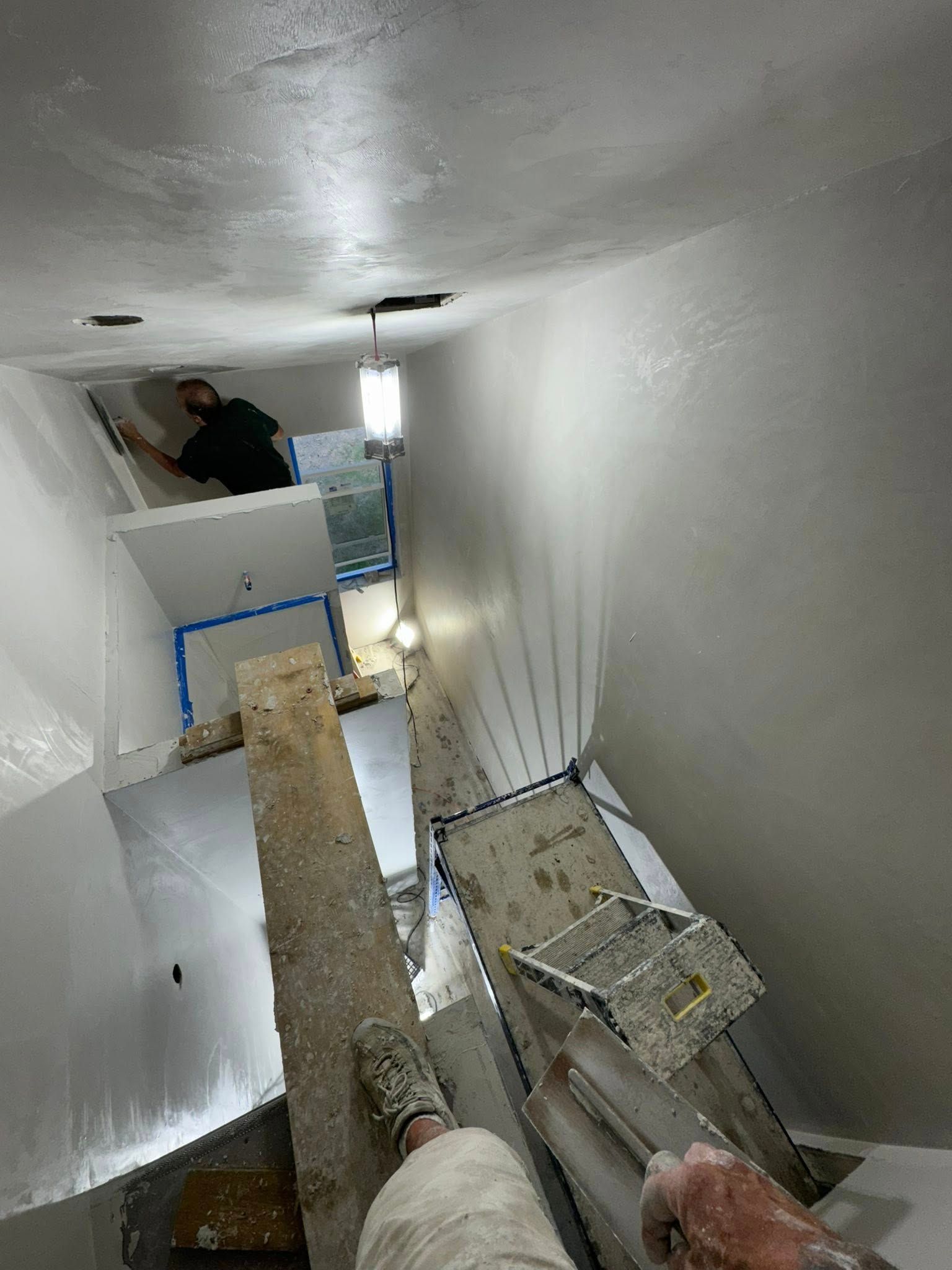 Person applies coating to white walls in a narrow space with scaffolding and overhead lights. Person applies coating to white walls in a narrow space with scaffolding and overhead lights.