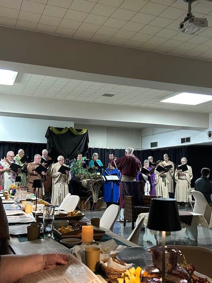 Choir performance in a banquet hall, with singers in beige robes around a conductor at center stage.