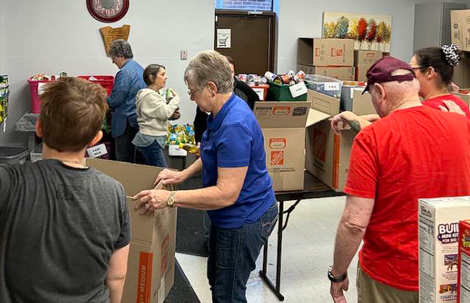 Volunteers packing boxes in a donation room with tables, supplies, and stacked cartons