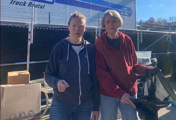 Two people standing beside a truck, one in a blue hoodie and one in a red jacket, holding items outdoors.