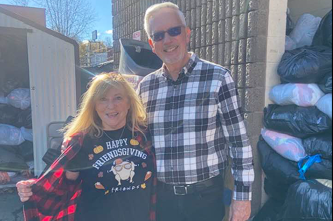 Two people smiling outdoors beside stacked bags and a shed, wearing a plaid shirt and a black sweatshirt.