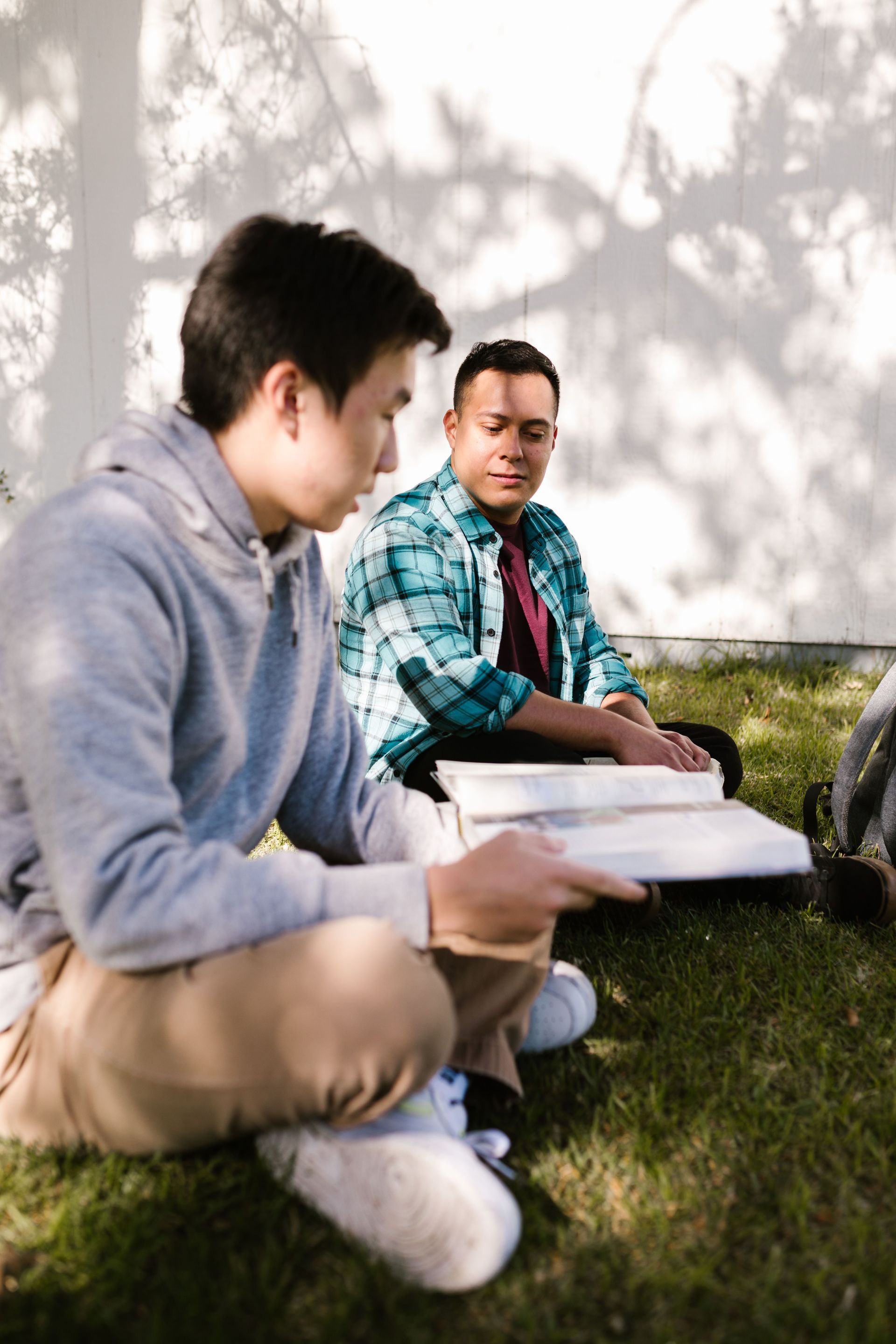 Two students sit on grass outdoors, studying with open books in a shaded area.
