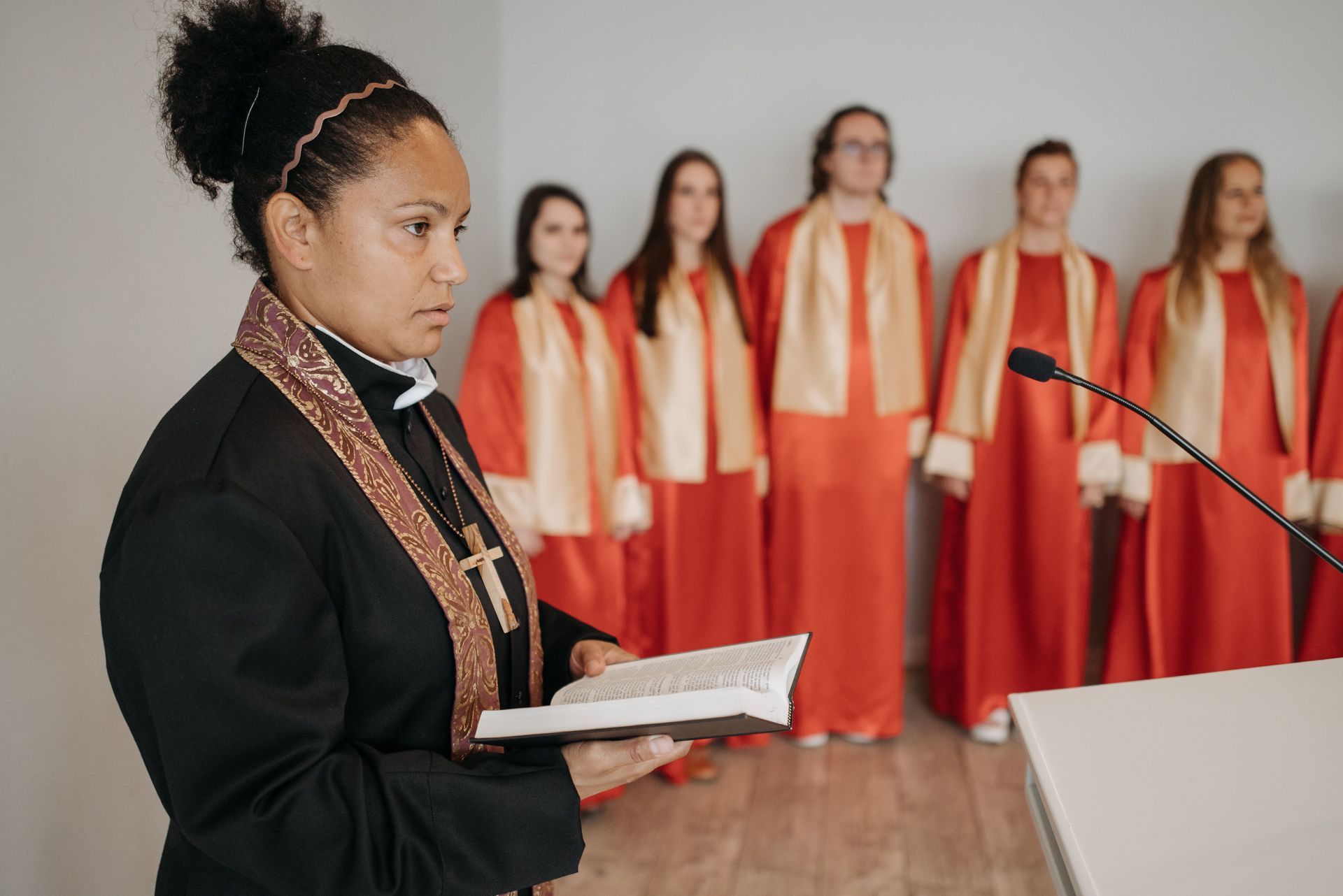 Woman reading from a book at a podium with a choir in red robes behind her