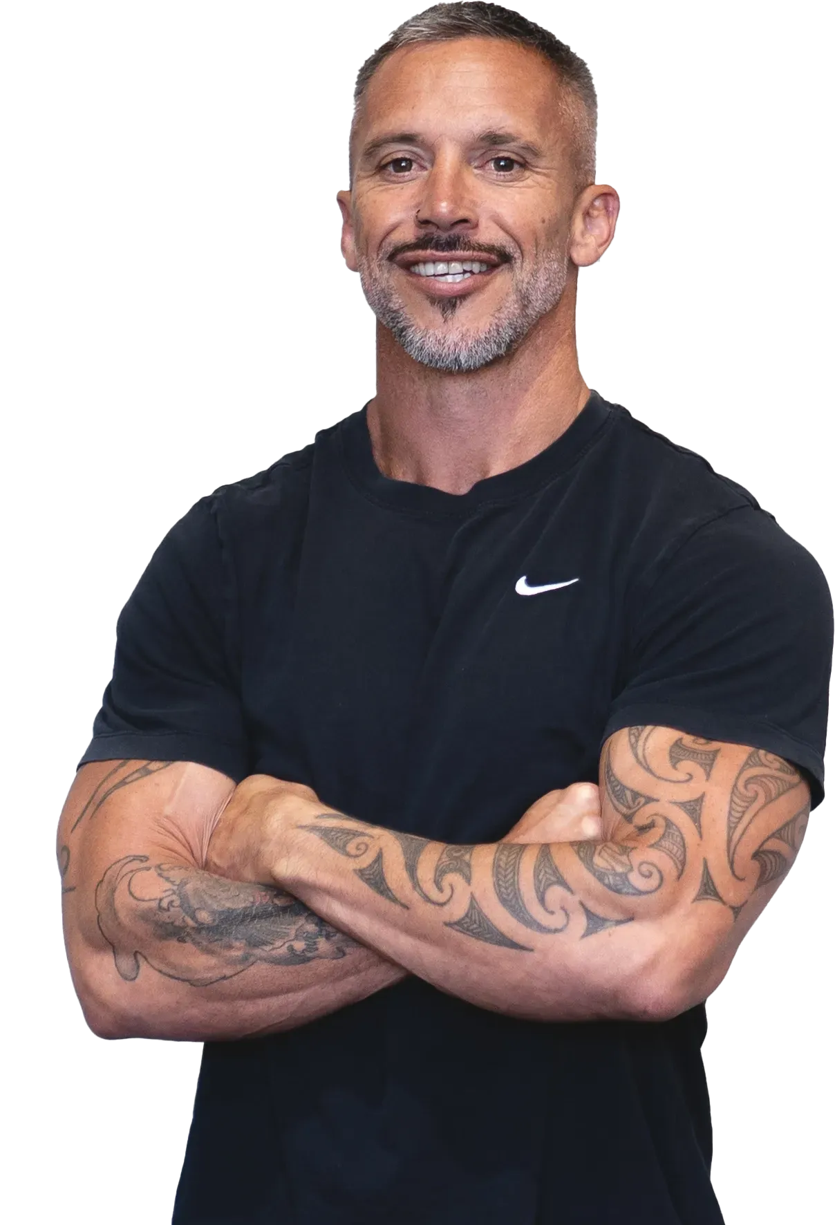 A man with a beard and tattoos is standing with his arms crossed and smiling. TBW FITNESS