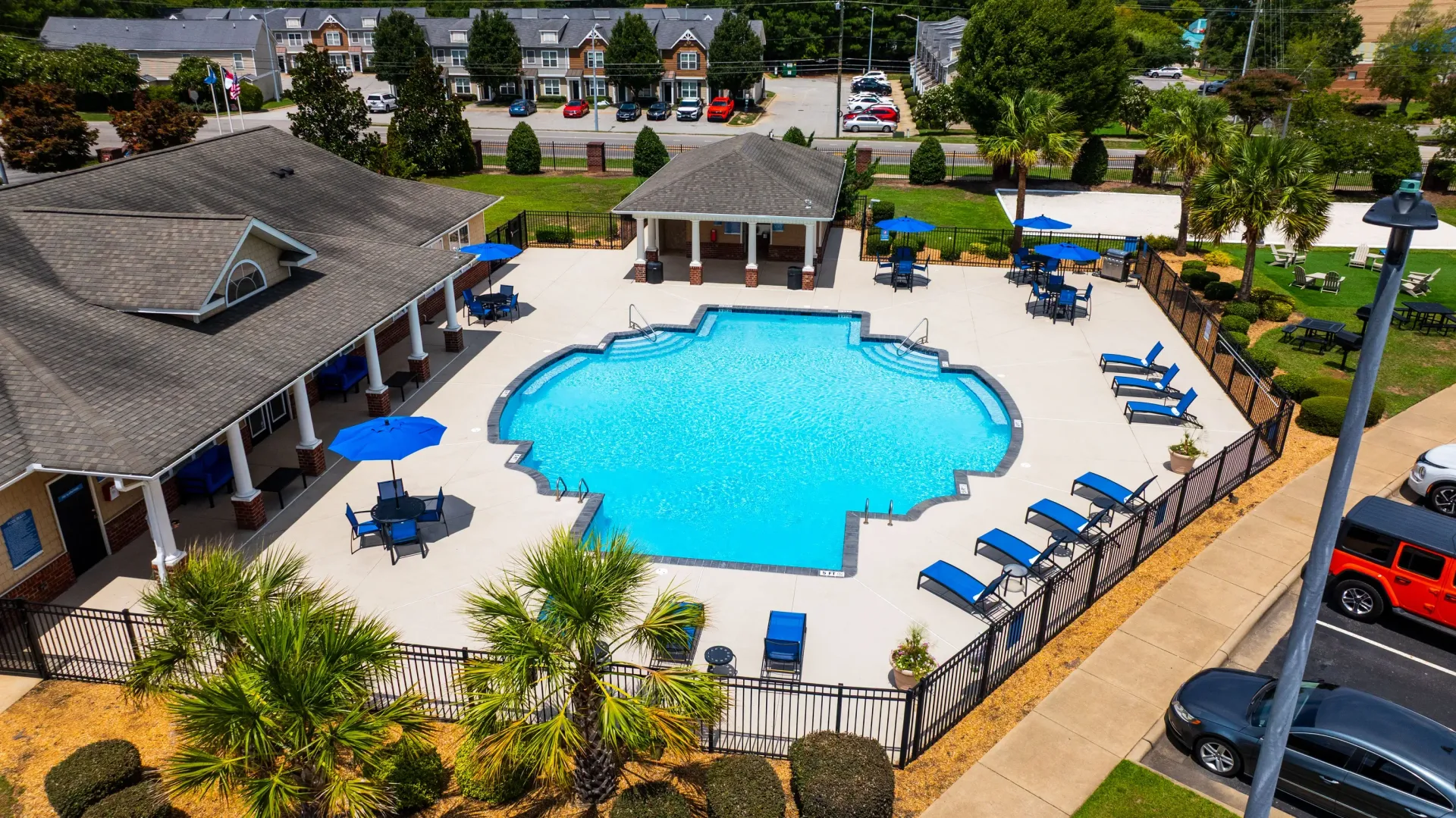 Aerial view of a large, freeform swimming pool surrounded by lounge chairs and umbrellas, with a covered pavilion and apartment buildings in the background.