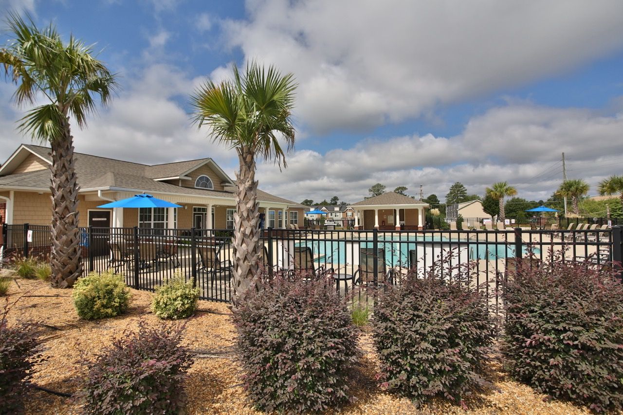 Outdoor apartment community pool with palm trees, fencing, and blue umbrellas.