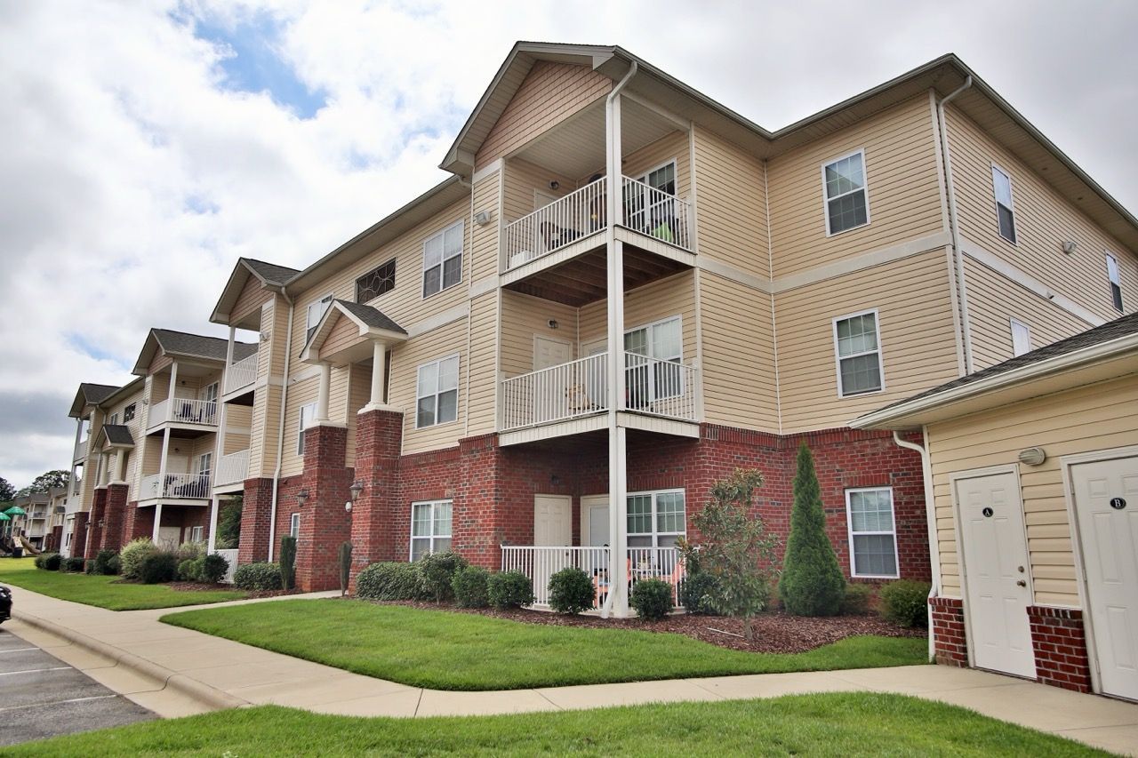 Exterior view of a beige, multi-unit apartment building with brick base, white balconies, and landscaped lawn.