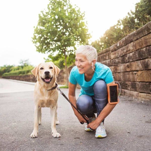 A person with short light hair in a teal athletic shirt crouches on a path to smile at a smiling yellow Labrador retriever at The Heights at McArthur ParkApartments inFayetteville,NC.