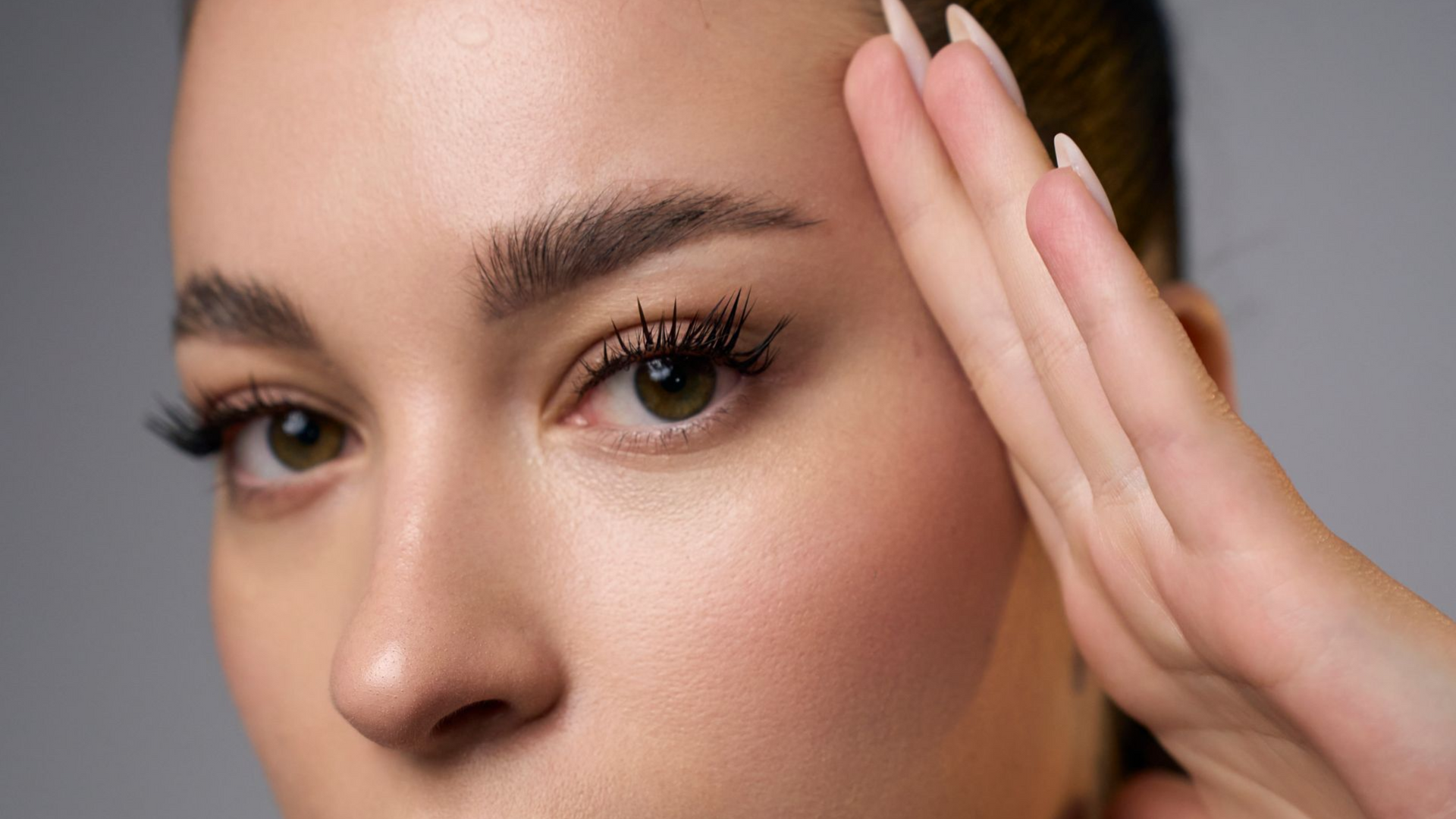 Close-up of a woman's face with long eyelashes and a hand near her temple against a neutral background