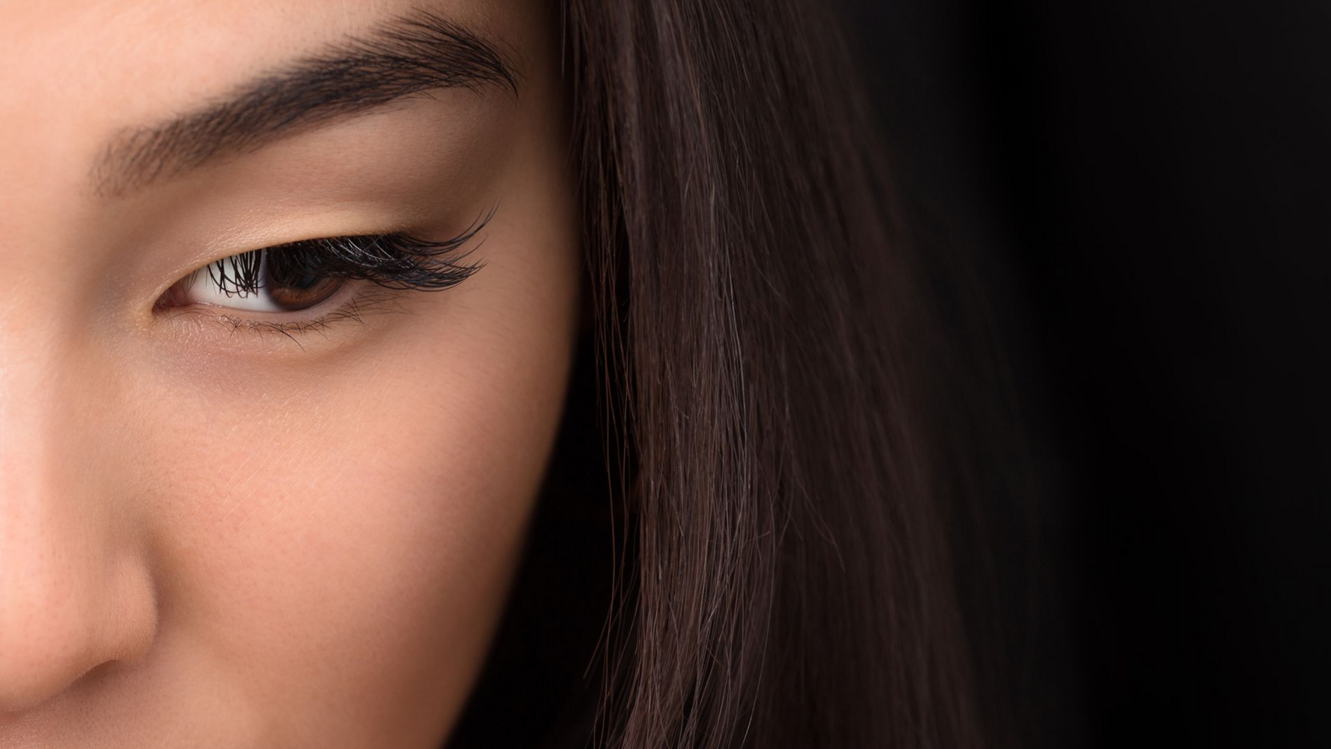 Close-up of a woman’s eye with long lashes and dark hair against a black background