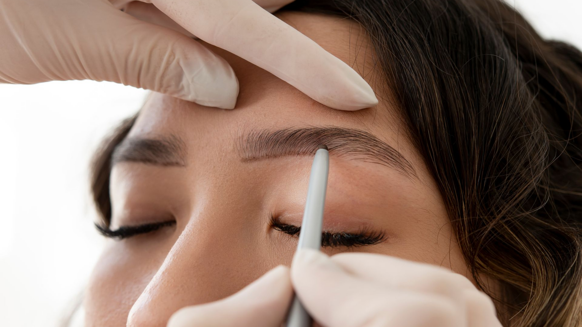 Cosmetologist shaping eyebrows with a brow pencil while holding the skin taut close-up