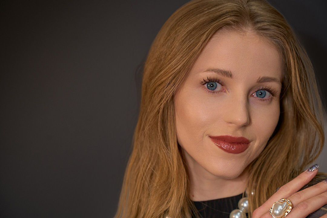 Portrait of a woman with long light-brown hair, blue eyes, and dark lipstick, wearing a pearl necklace and ornate ring.