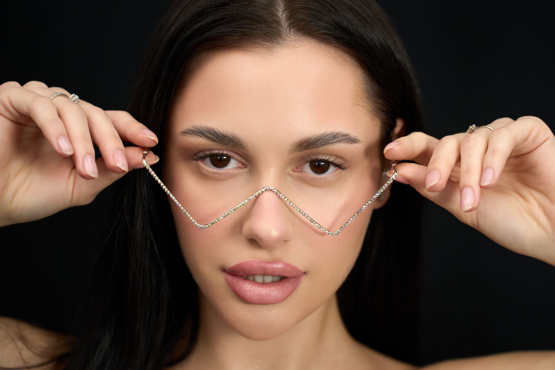 A person with long dark hair holds a silver chain necklace in a V-shape across the bridge of their nose against black.