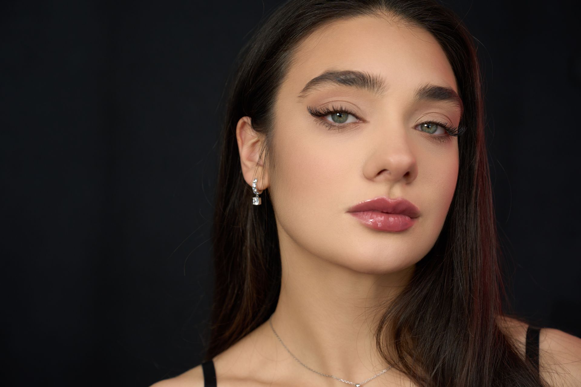 A person with dark hair and a neutral expression against a plain black background, wearing simple jewelry.