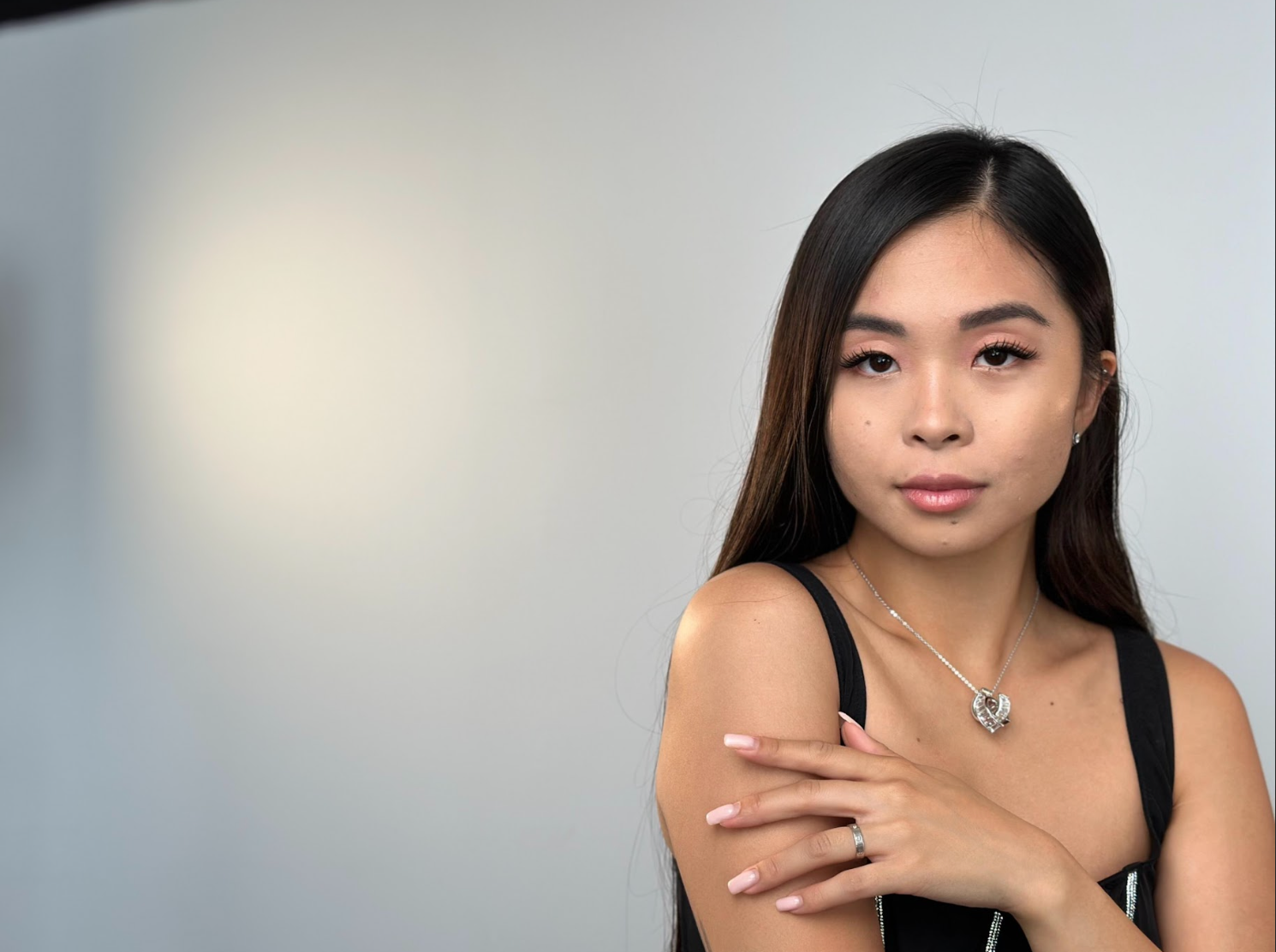 A woman wearing a necklace and a ring is standing in front of a white wall.