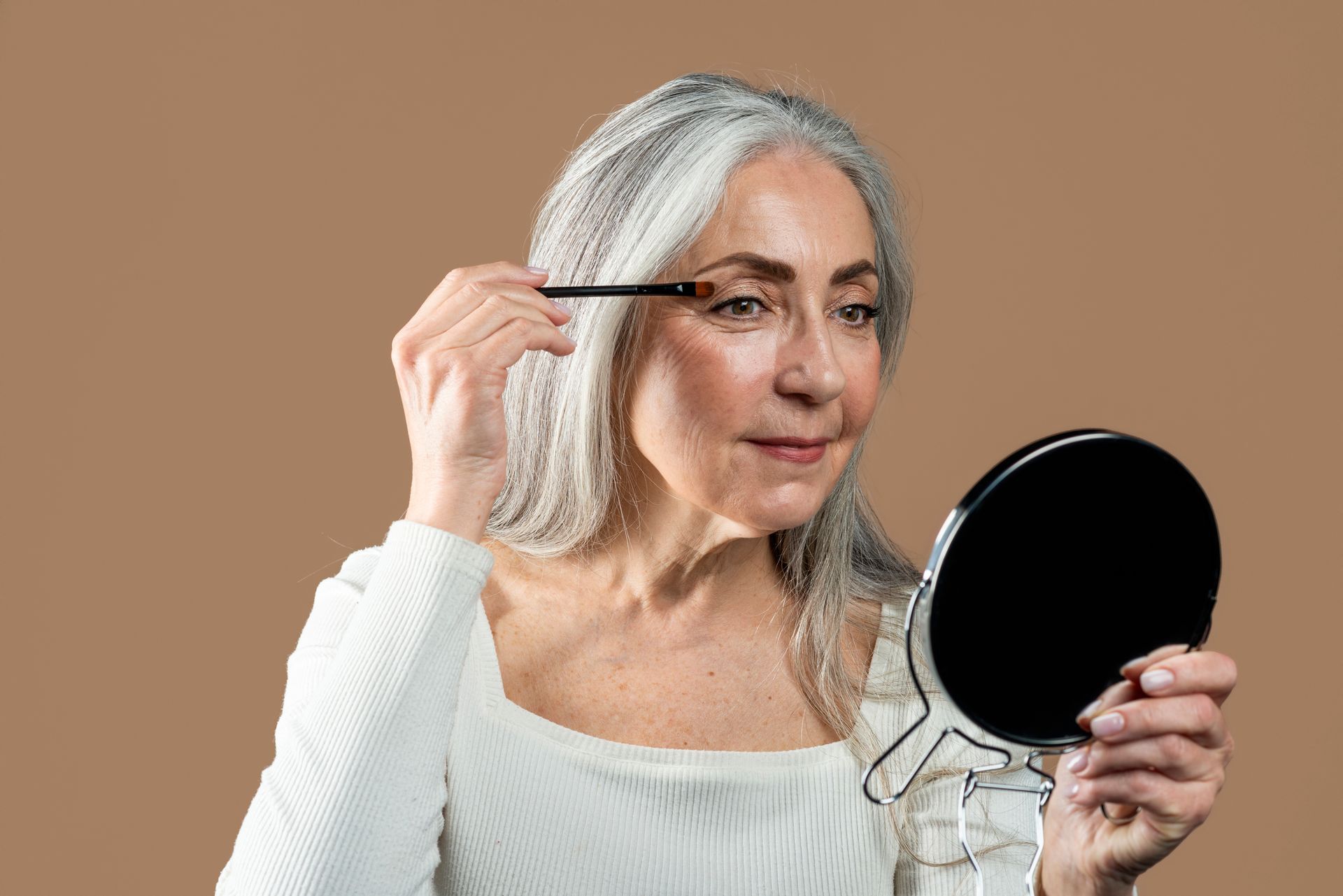 A person with long gray hair looking into a hand mirror while applying makeup to their eyelid against a brown background.