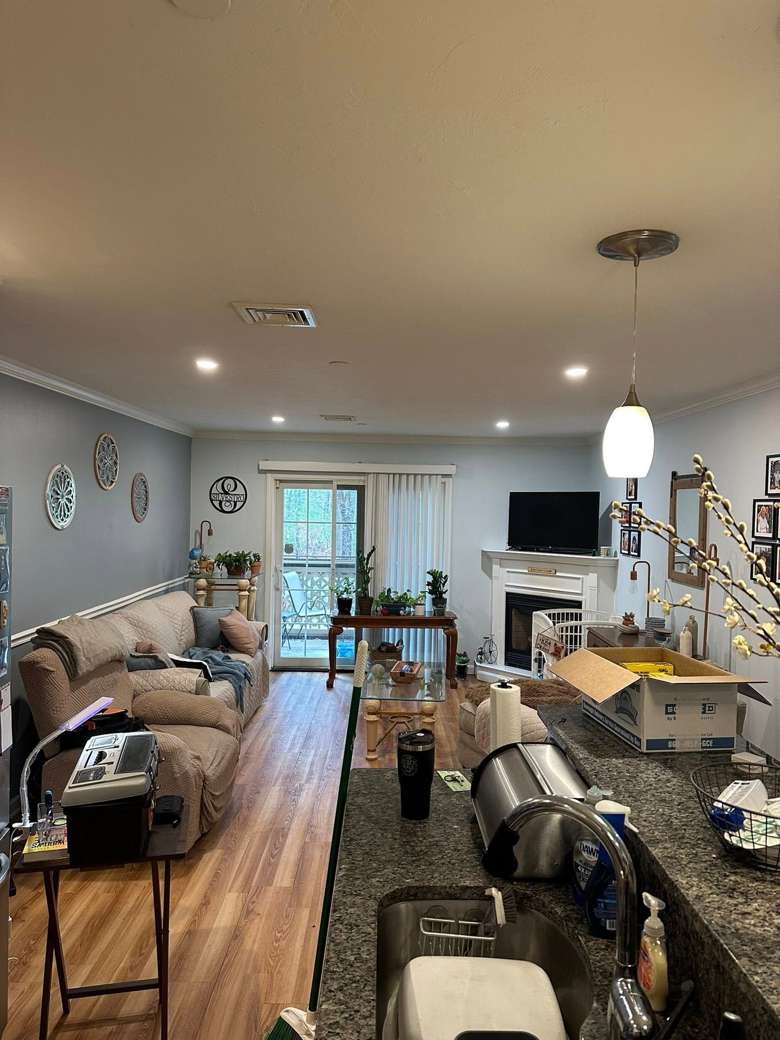 A kitchen with granite counters overlooks a living room with a beige sofa, sliding glass doors, and a TV on a fireplace.
