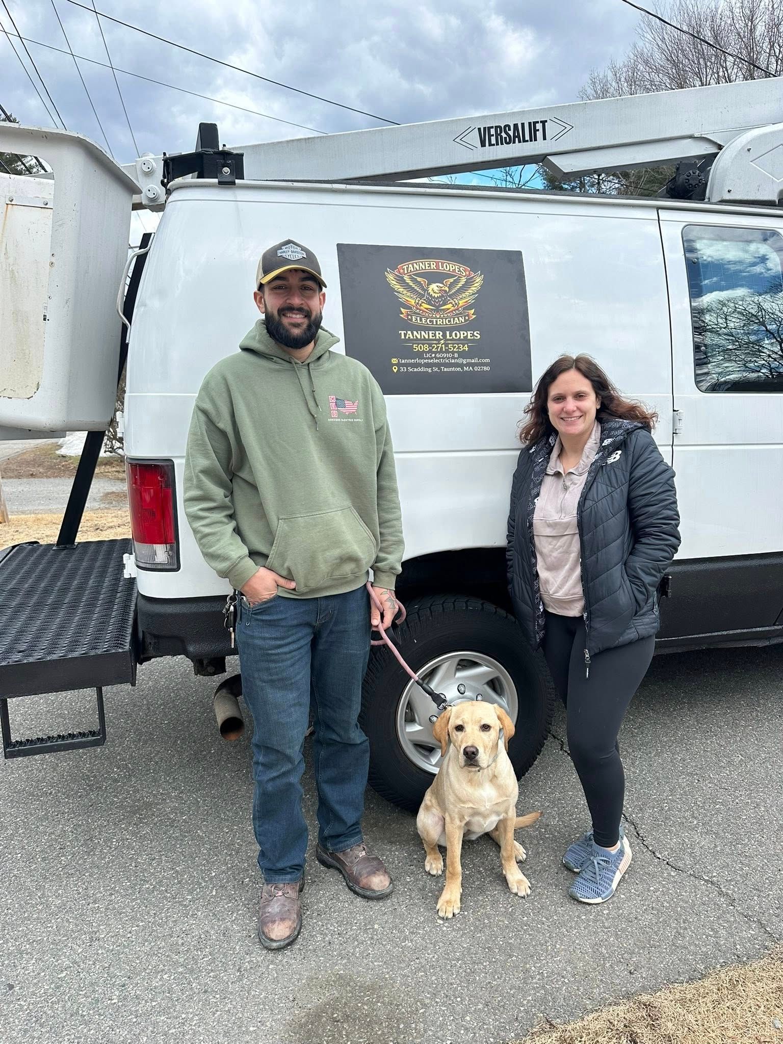 A person in a hoodie and a person in a jacket stand with a yellow Labrador retriever in front of a white utility van.