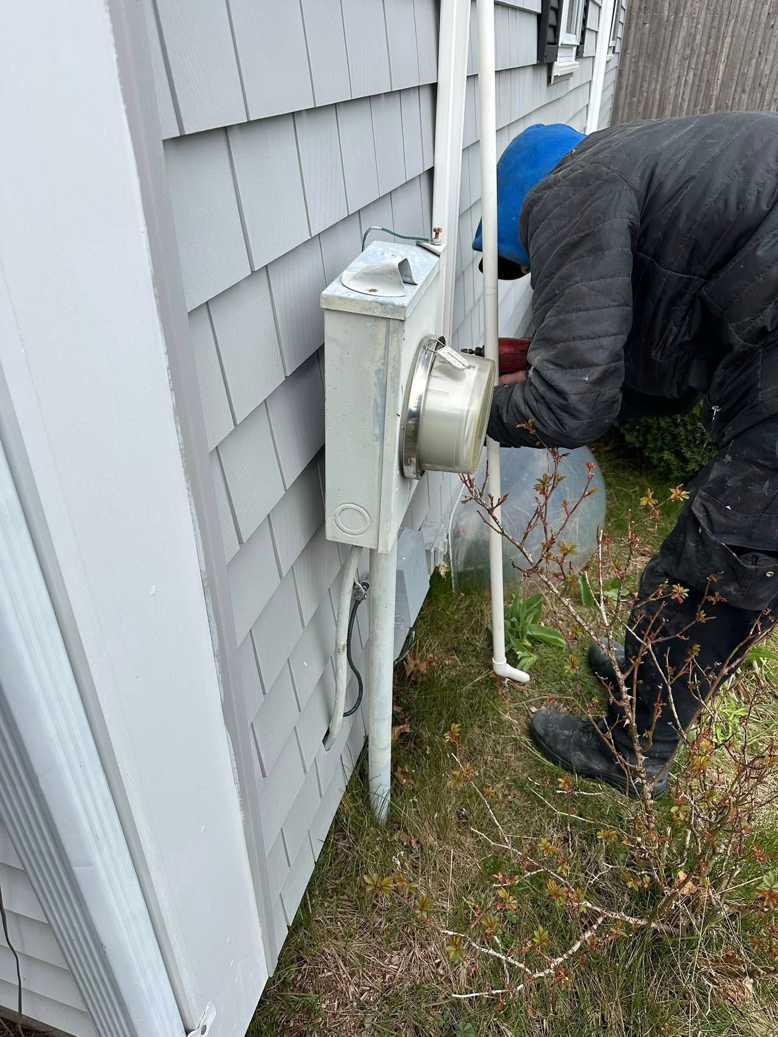 A person in a dark jacket and blue hood works on an electrical meter box attached to a light gray house wall.