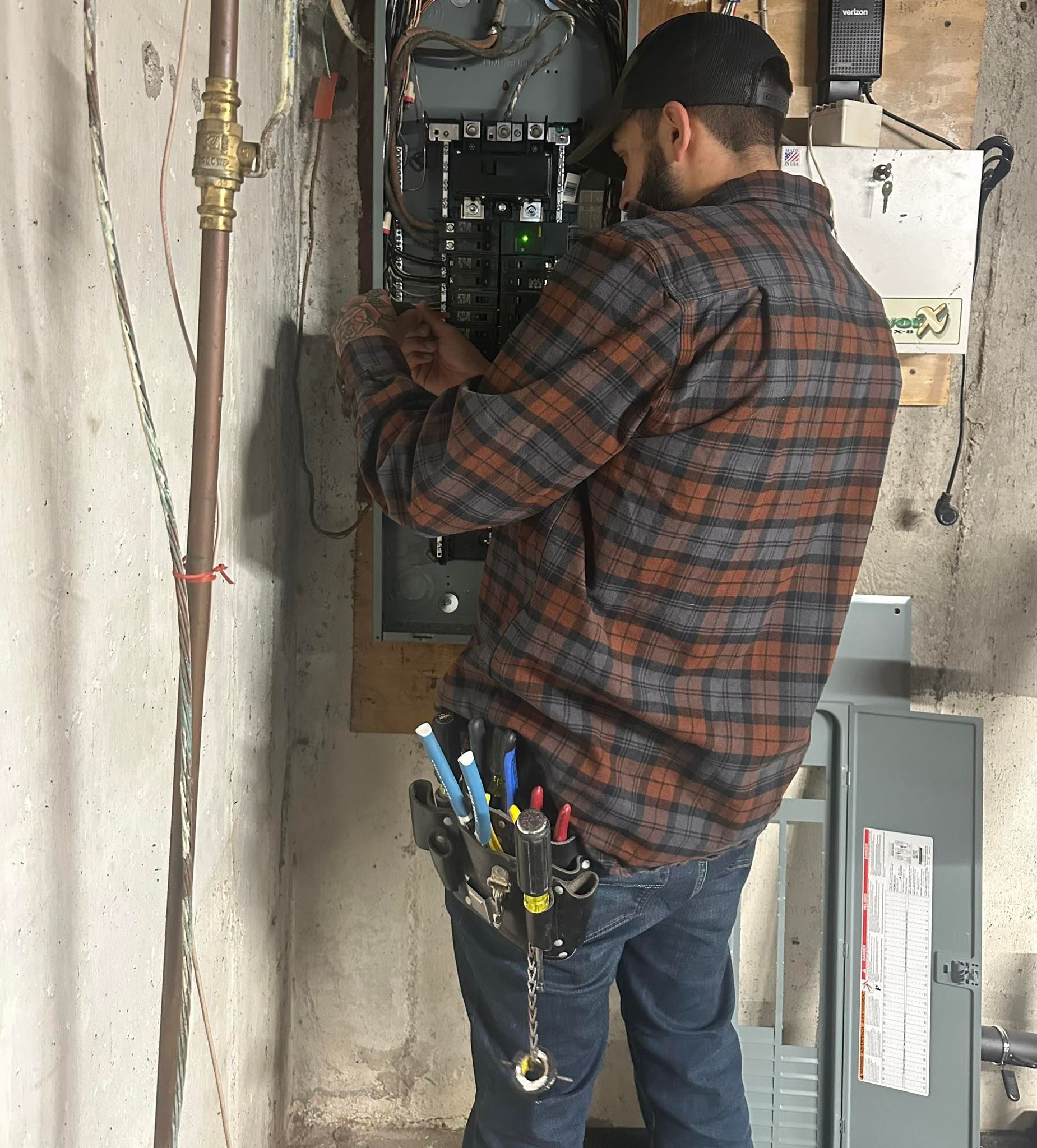 An electrician wearing a plaid shirt and tool belt works on an open electrical panel mounted to a concrete wall.