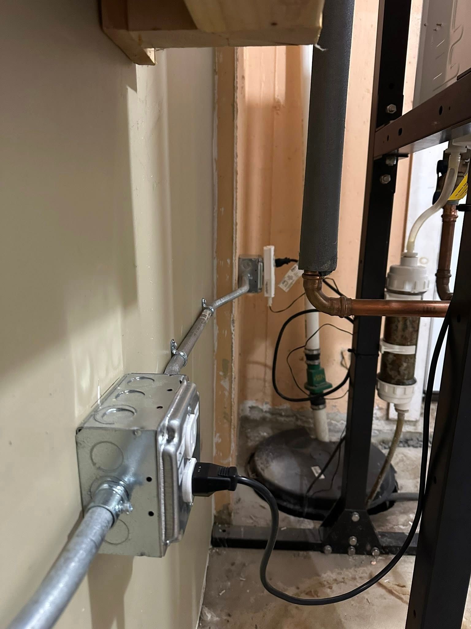 Electrical metal conduit and outlet box mounted on a wall with a plugged-in cord near a sump pump and water filter system.