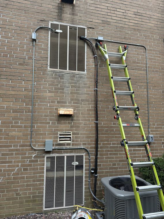 A tall extension ladder leans against a brick wall next to HVAC vents, exposed electrical conduits, and a compressor unit.