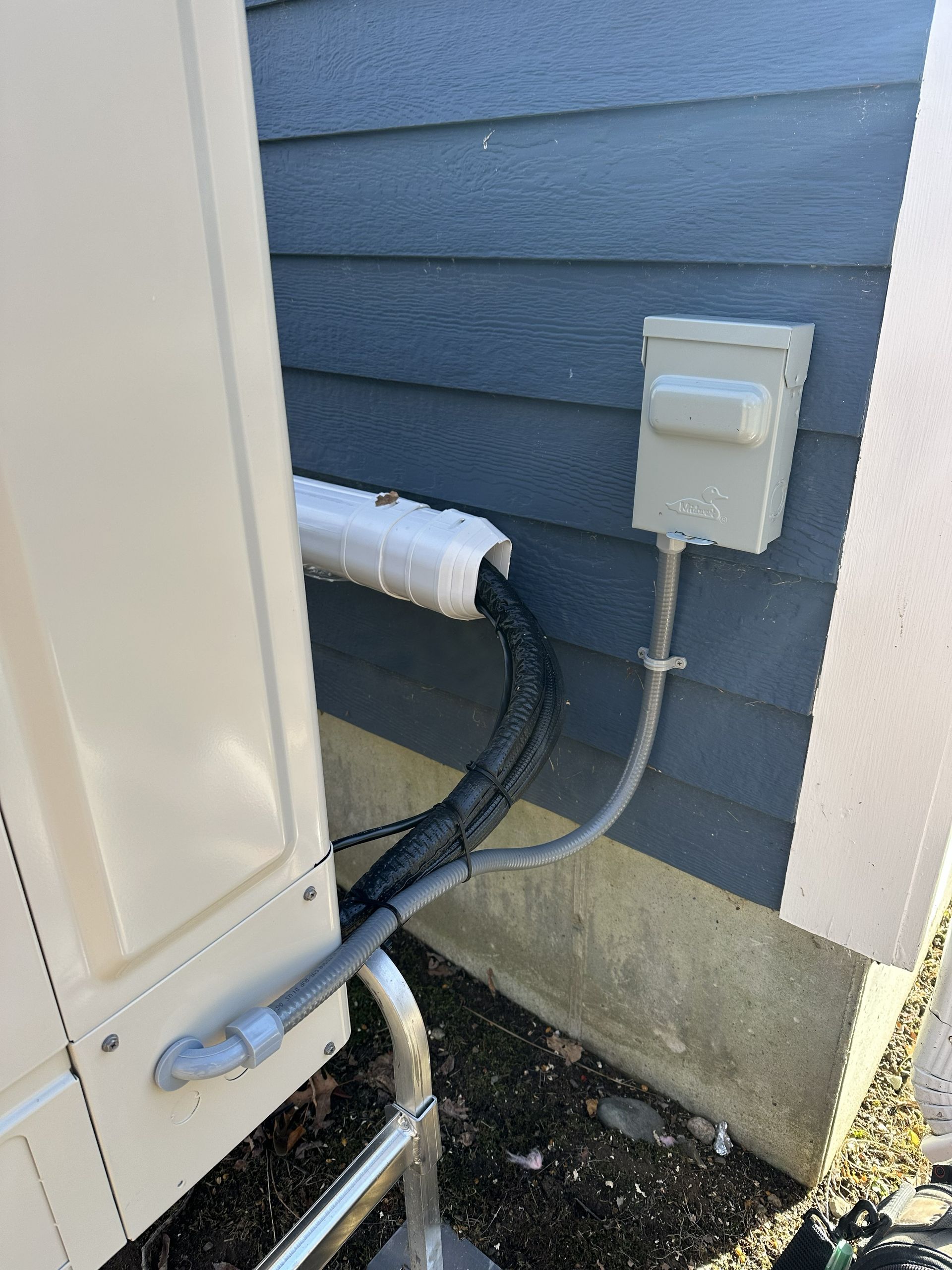 Outdoor HVAC unit connected to a gray exterior wall with a white insulated pipe and an electrical disconnect box.
