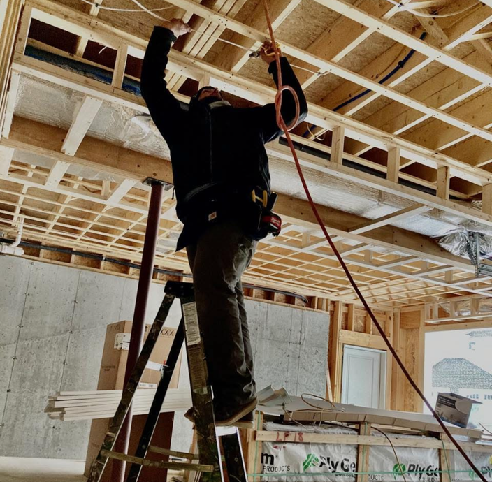 A person in dark clothing stands on a stepladder, reaching up to install electrical wiring in an unfinished wood building.