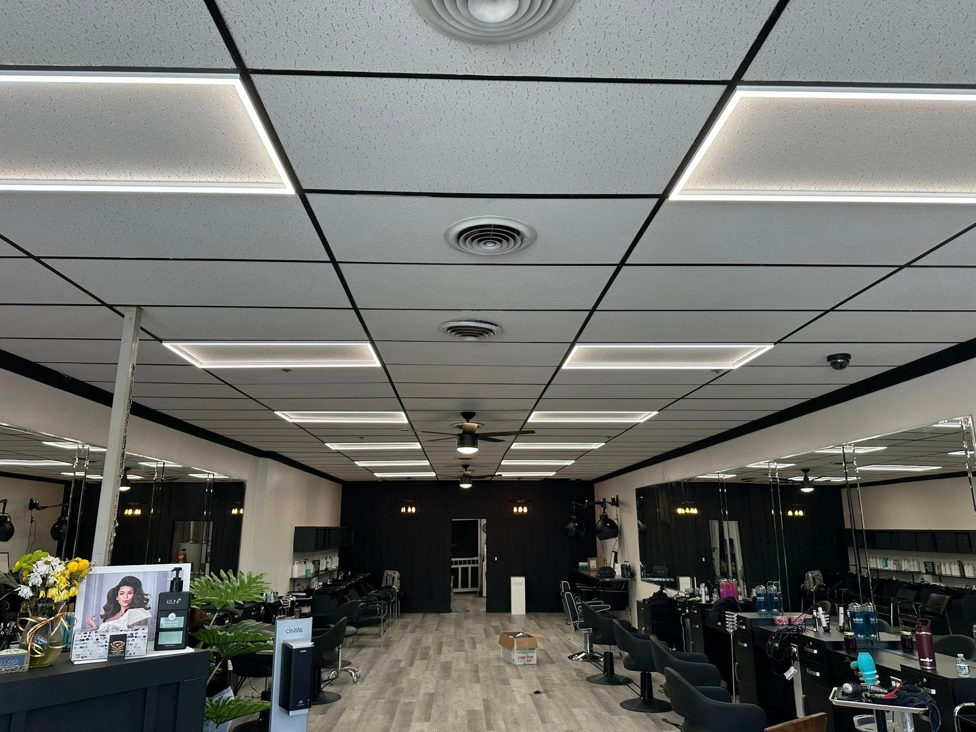 Inside a hair salon with a drop ceiling, lighting panels, and rows of black salon chairs on a light wood floor.