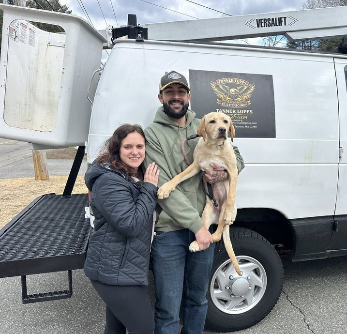 A couple smiling while holding a light-colored dog in front of a white work van with a bucket lift.