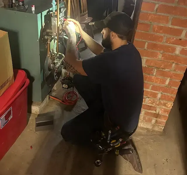 A person with a beard and baseball cap crouches while using a multimeter to inspect the components of a green boiler.