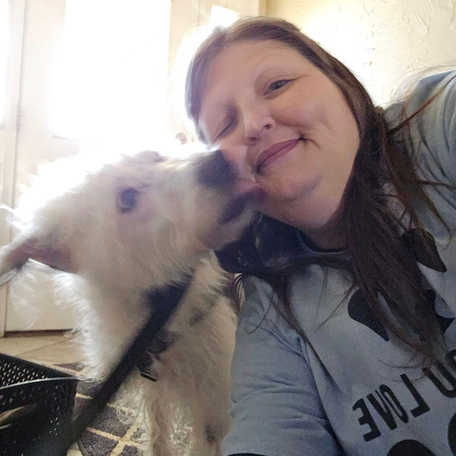 Woman smiles as white dog licks her cheek. They are indoors.