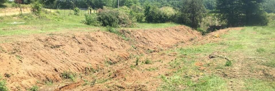 Dry grassy field with a large exposed dirt patch and trees in the background