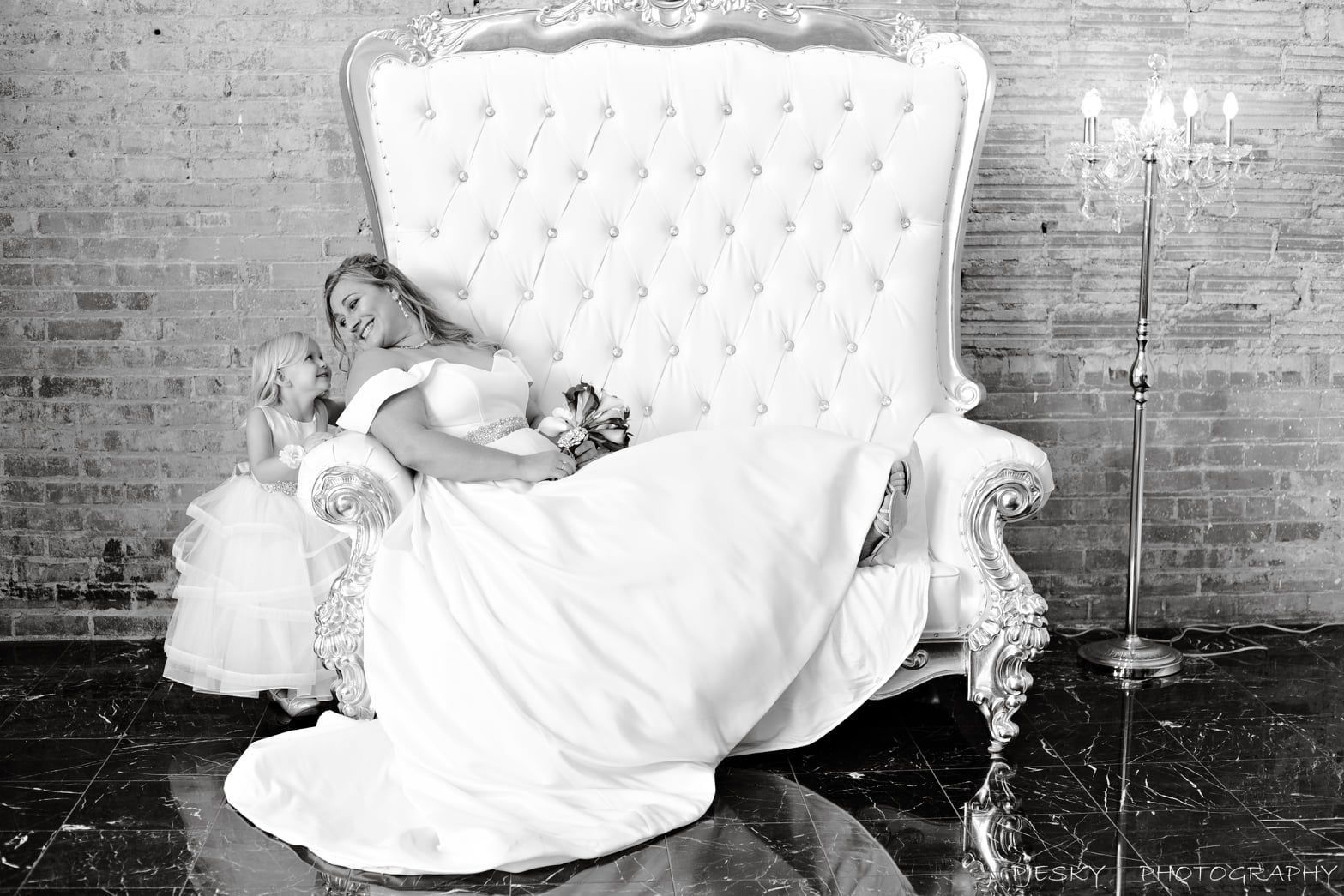 A black and white photo of a bride and her flower girl sitting on a couch.