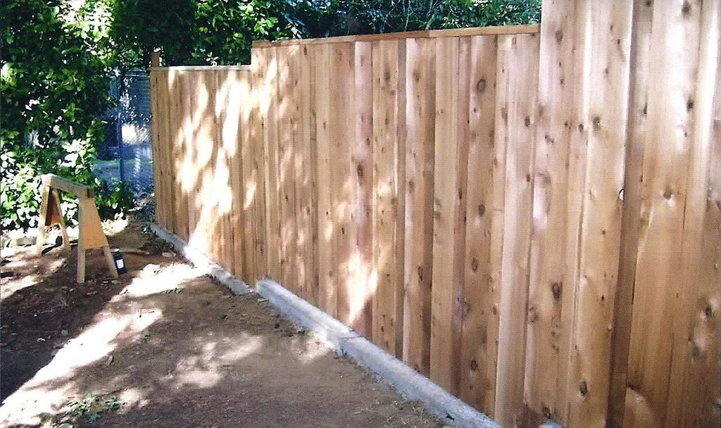 Wooden fence in a yard with dirt and a concrete base. Sunlight casts shadows on the fence.