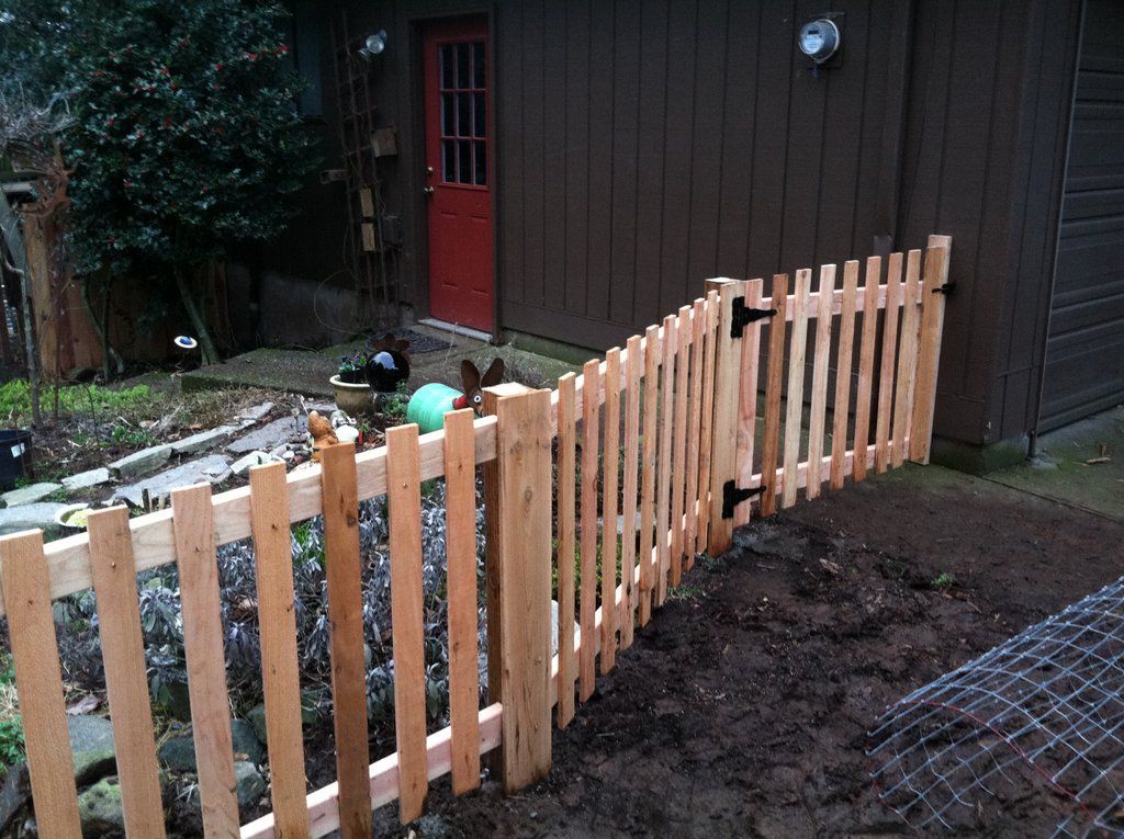 Wooden picket fence with gate in front of a brown building and red door.