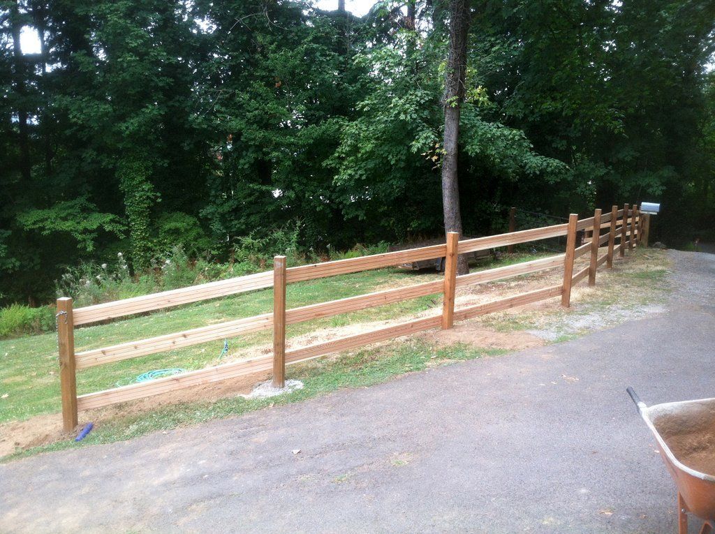 Brown split-rail fence alongside a paved driveway and green foliage. A wheelbarrow is in the foreground.