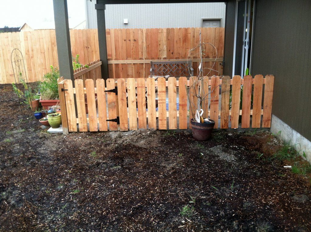 Wooden picket fence within a backyard, with taller wooden fence in the background, on a gravel ground.