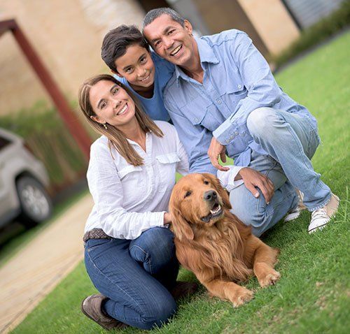 A family of three smiling with a golden retriever on a grassy lawn in front of a house.