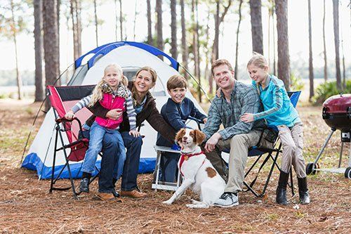 A family with a dog poses in front of a camping tent in a wooded area, with a grill visible in the background.