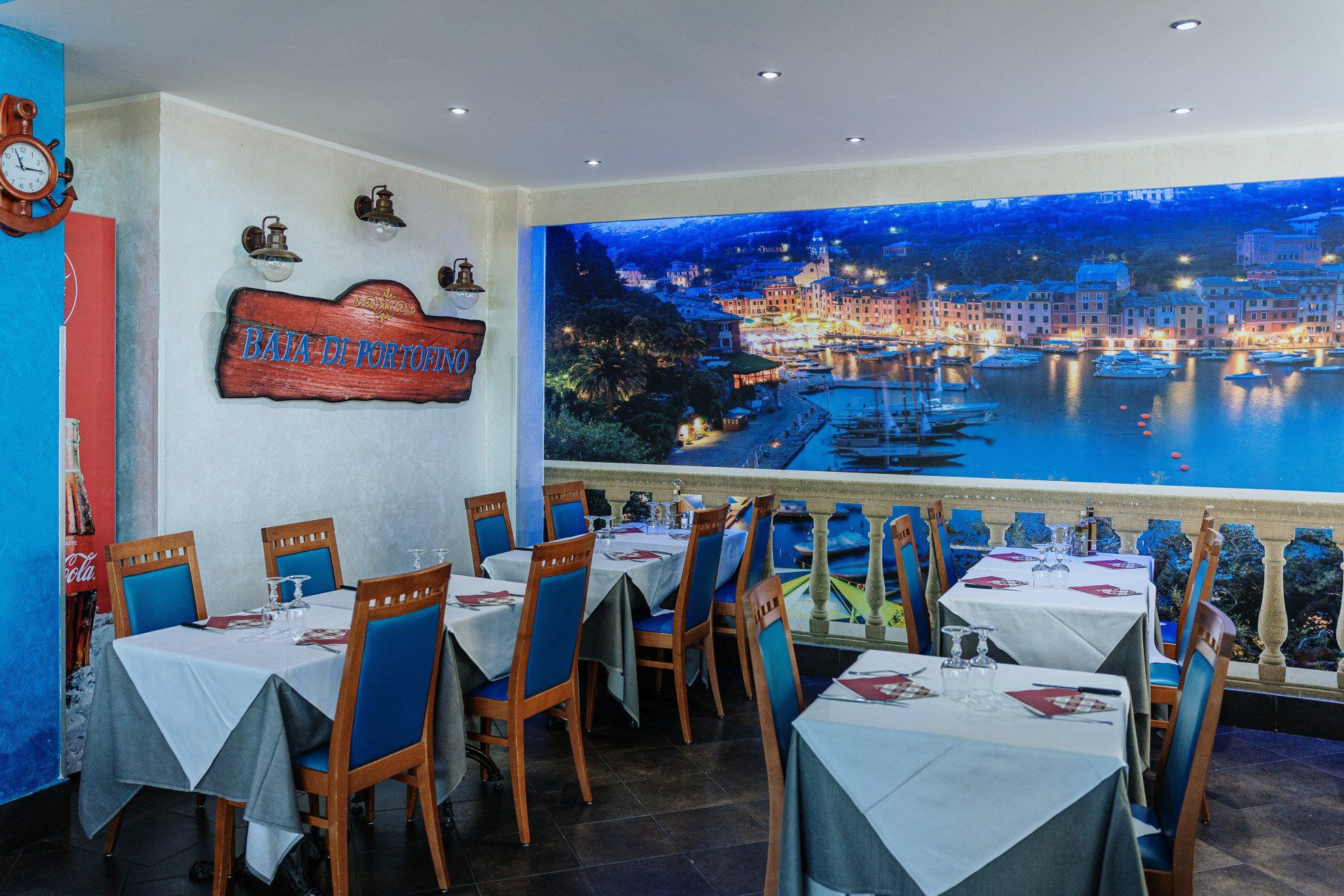 Interno del ristorante con tavoli apparecchiati per cenare, con vista su un murale raffigurante un porto di notte.