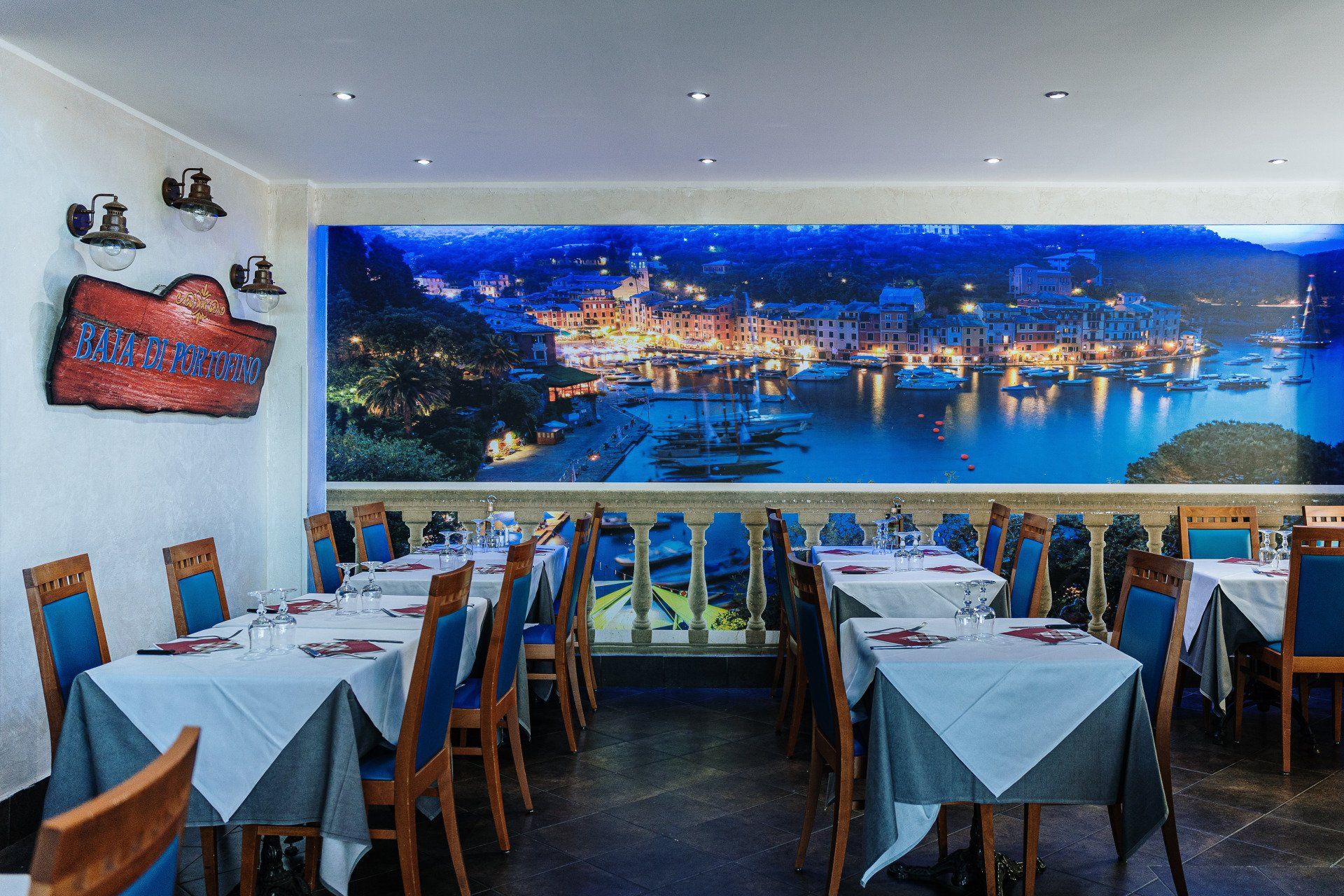 Interno di un ristorante con tavoli apparecchiati e un grande murale raffigurante una città costiera.