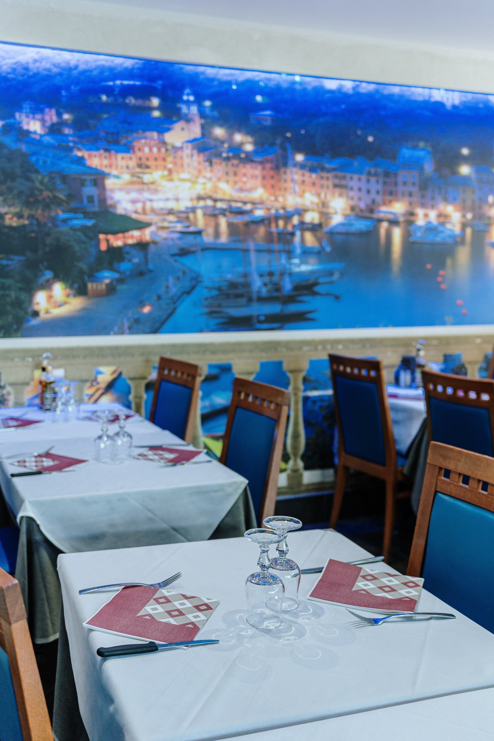 Tavoli del ristorante apparecchiati per cenare, con sedie blu e un murale raffigurante una scena portuale sullo sfondo.