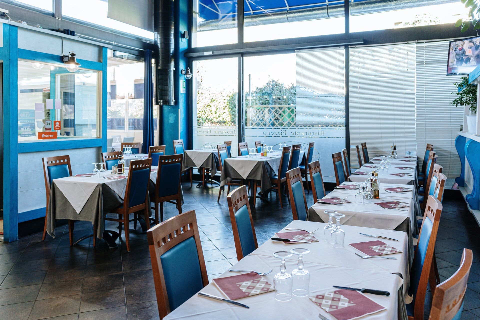Interno del ristorante con tavoli apparecchiati per la cena, sedie e dettagli blu. Ampie finestre lasciano entrare la luce naturale.