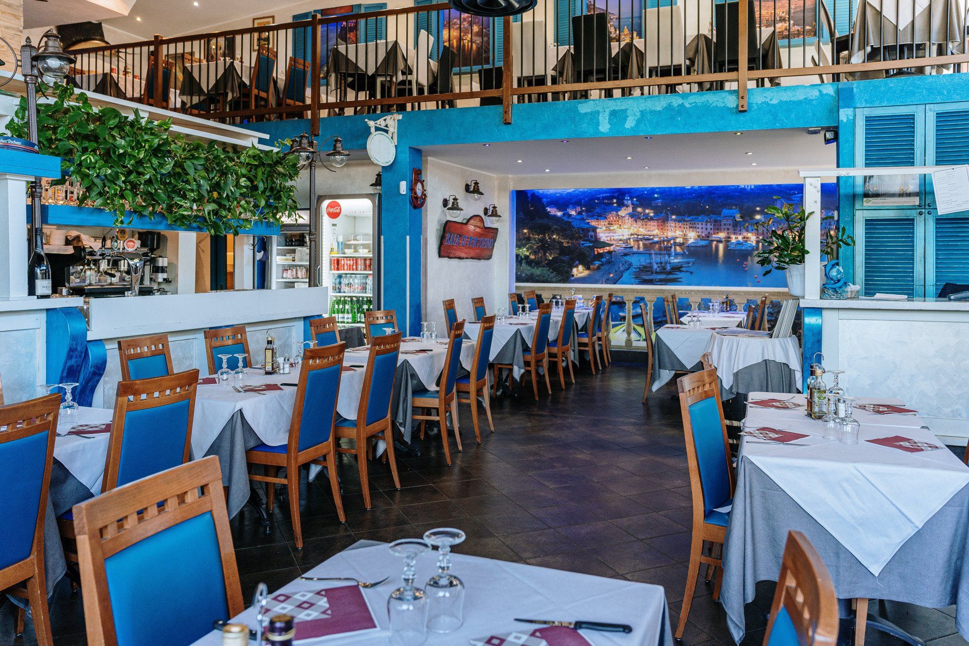 Interno del ristorante: tavoli apparecchiati con tovaglie bianche, sedie blu, murale di una città costiera sulla parete di fondo, balcone in alto.