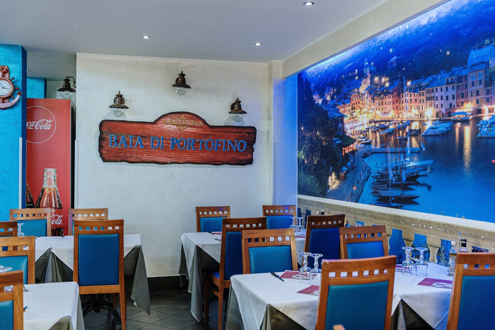 Interno del ristorante con tavoli apparecchiati per cenare, decorazioni a tema nautico e un grande murale raffigurante una città costiera.