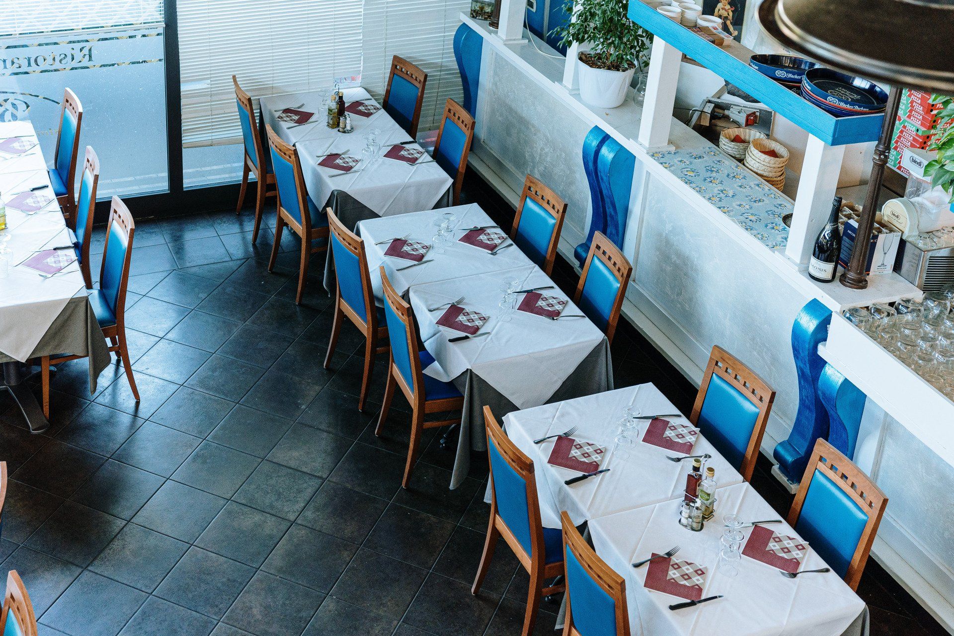 Interno del ristorante con decorazioni blu e bianche; tavoli apparecchiati con tovaglie e sedie bianche, vista dall'alto.