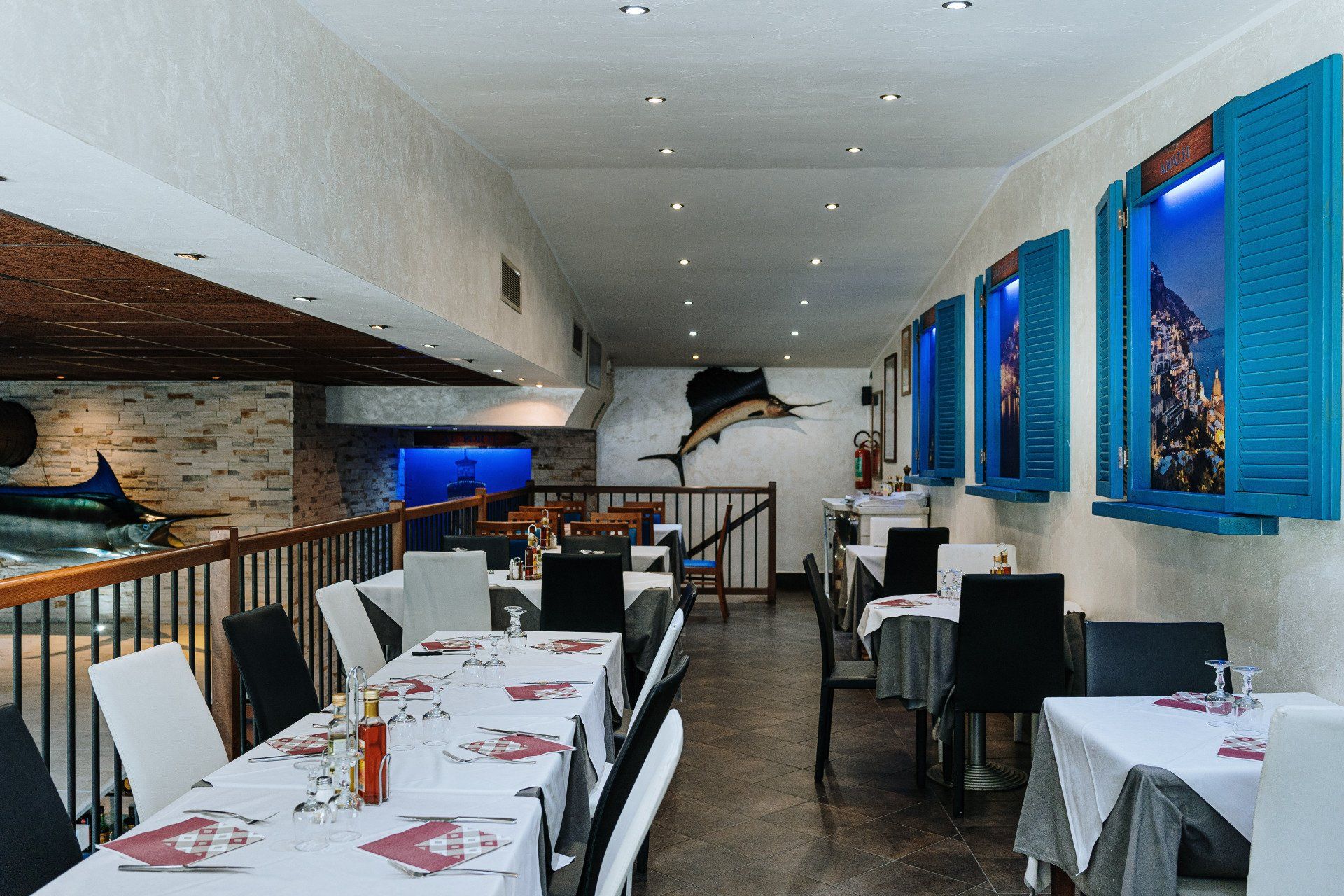 Interno di un ristorante con tavoli apparecchiati per cenare, persiane blu e decorazioni a forma di pesce.
